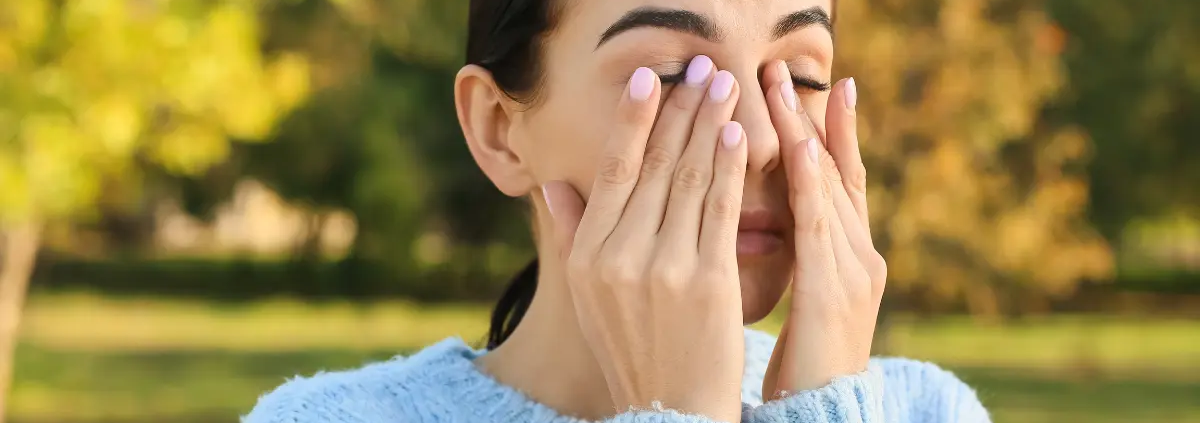Donna all’aperto che si strofina gli occhi per prurito dovuto ad allergie primaverili, con sfondo di alberi e erba.