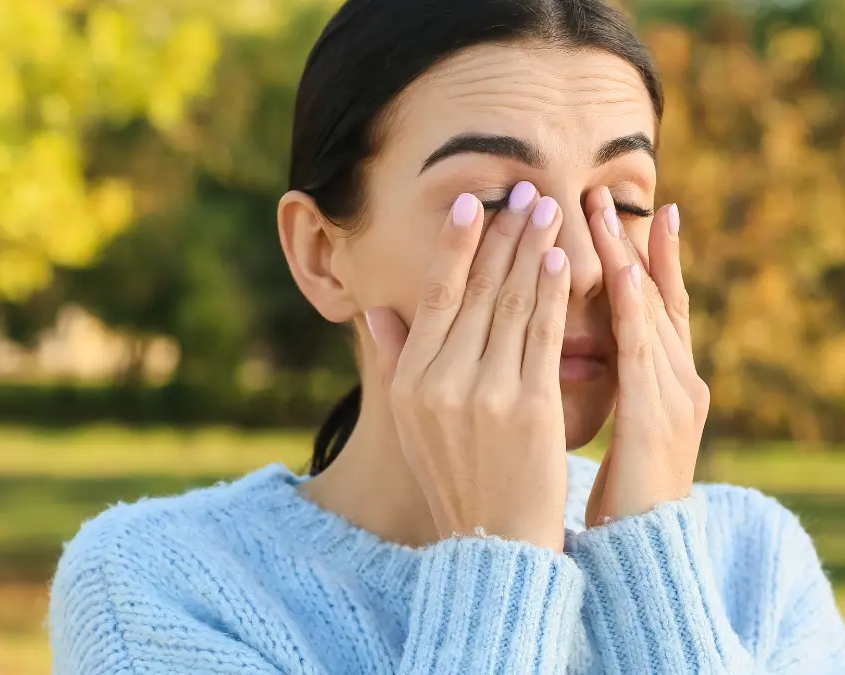 Donna all’aperto che si strofina gli occhi per prurito dovuto ad allergie primaverili, con sfondo di alberi e erba.
